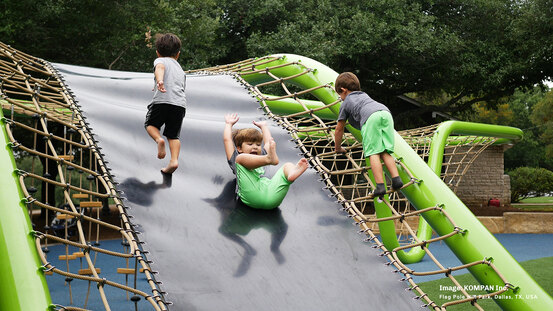 Three children play on a green playground structure with a central slide and climbing ropes at Flag Pole Hill Park in Dallas, Texas.