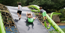 Three children play on a green playground structure with a central slide and climbing ropes at Flag Pole Hill Park in Dallas, Texas.